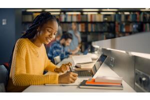 uma mulher universitária estudando usando um notebook em uma biblioteca. Ao fundo observamos algumas pessoas desfocadas.