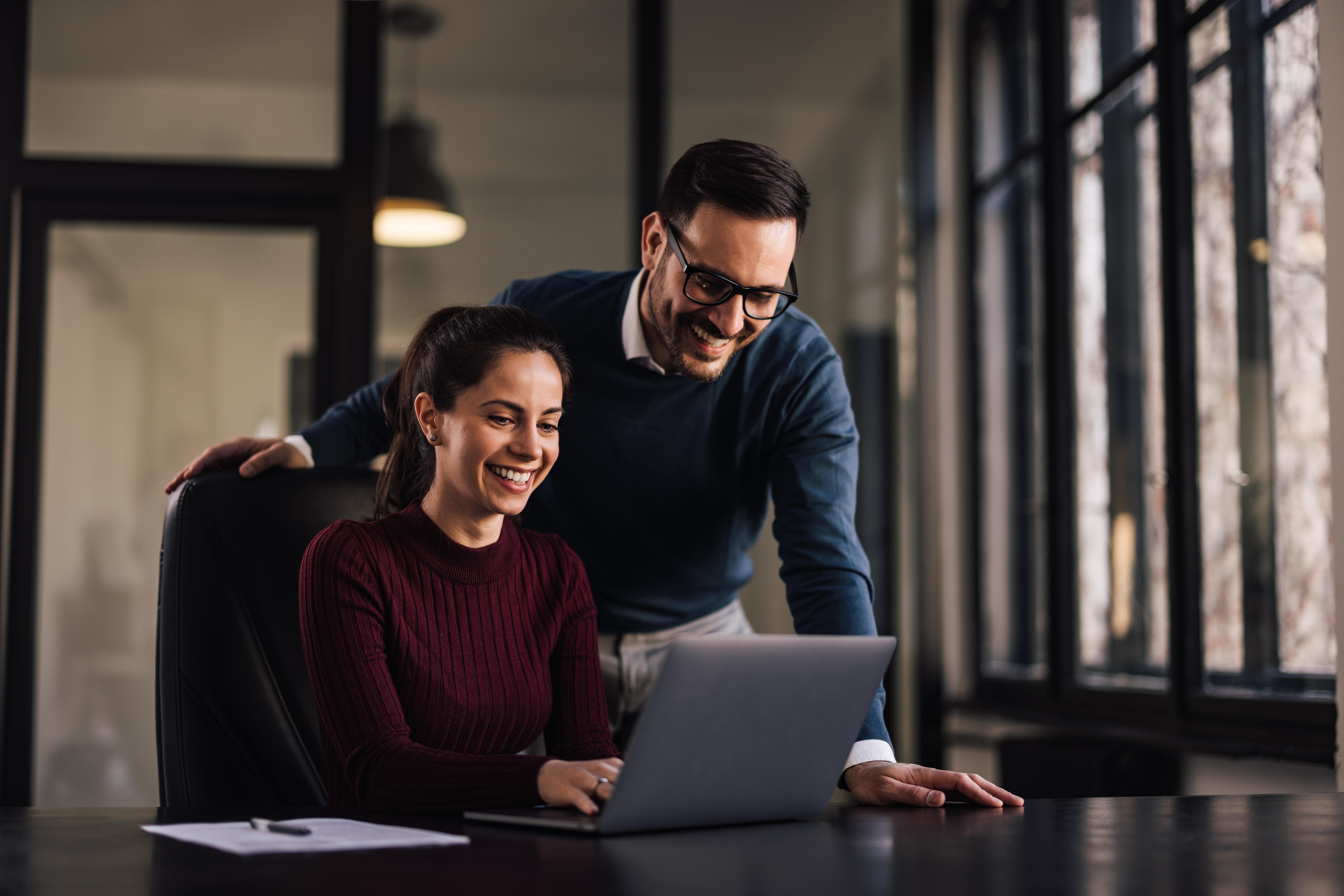Em um escritório, uma mulher digita em seu notebook, enquanto seu colega de trabalho está em pé ao seu lado, ambos sorriem.
