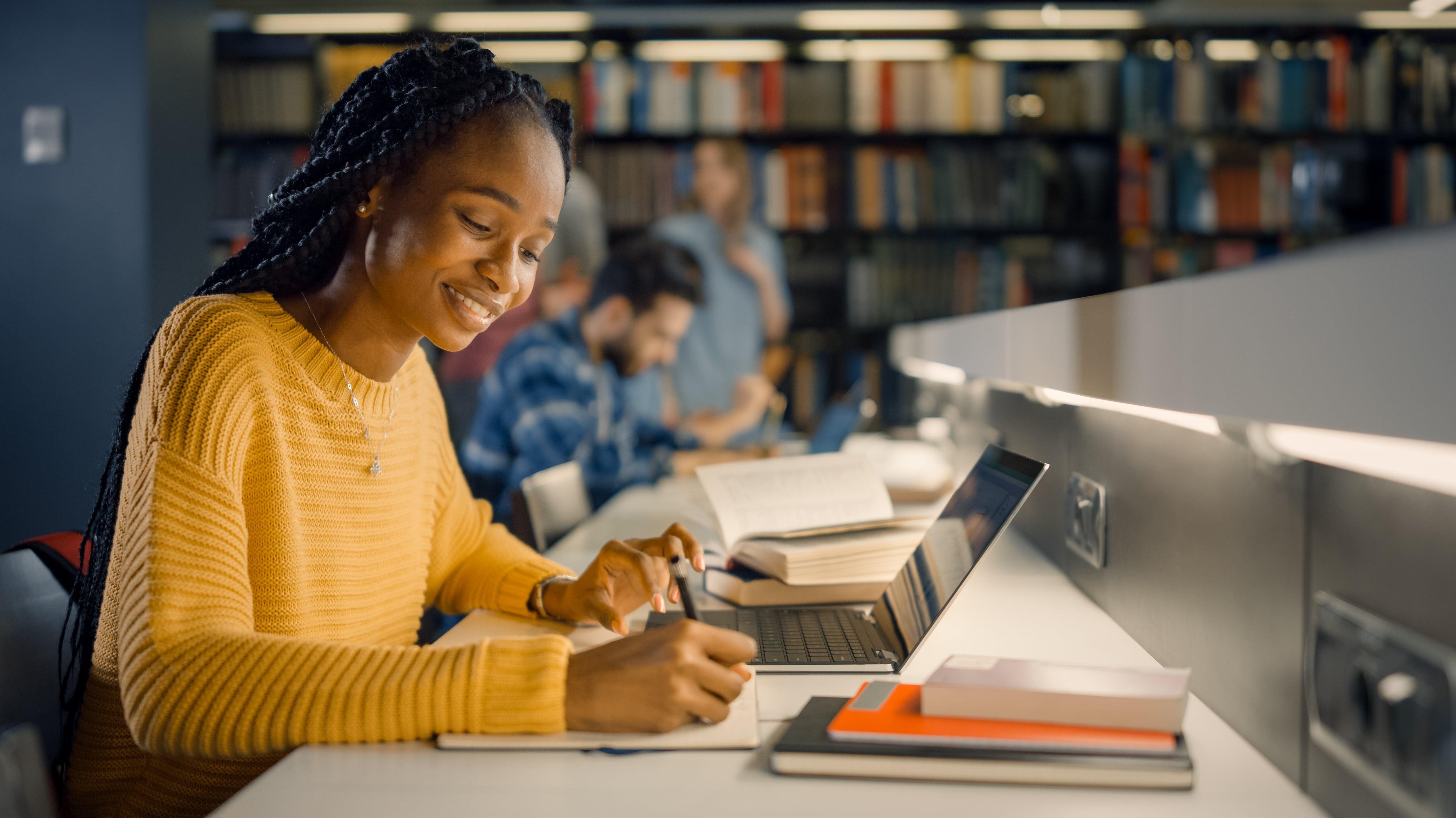 uma mulher universitária estudando usando um notebook em uma biblioteca. Ao fundo observamos algumas pessoas desfocadas.