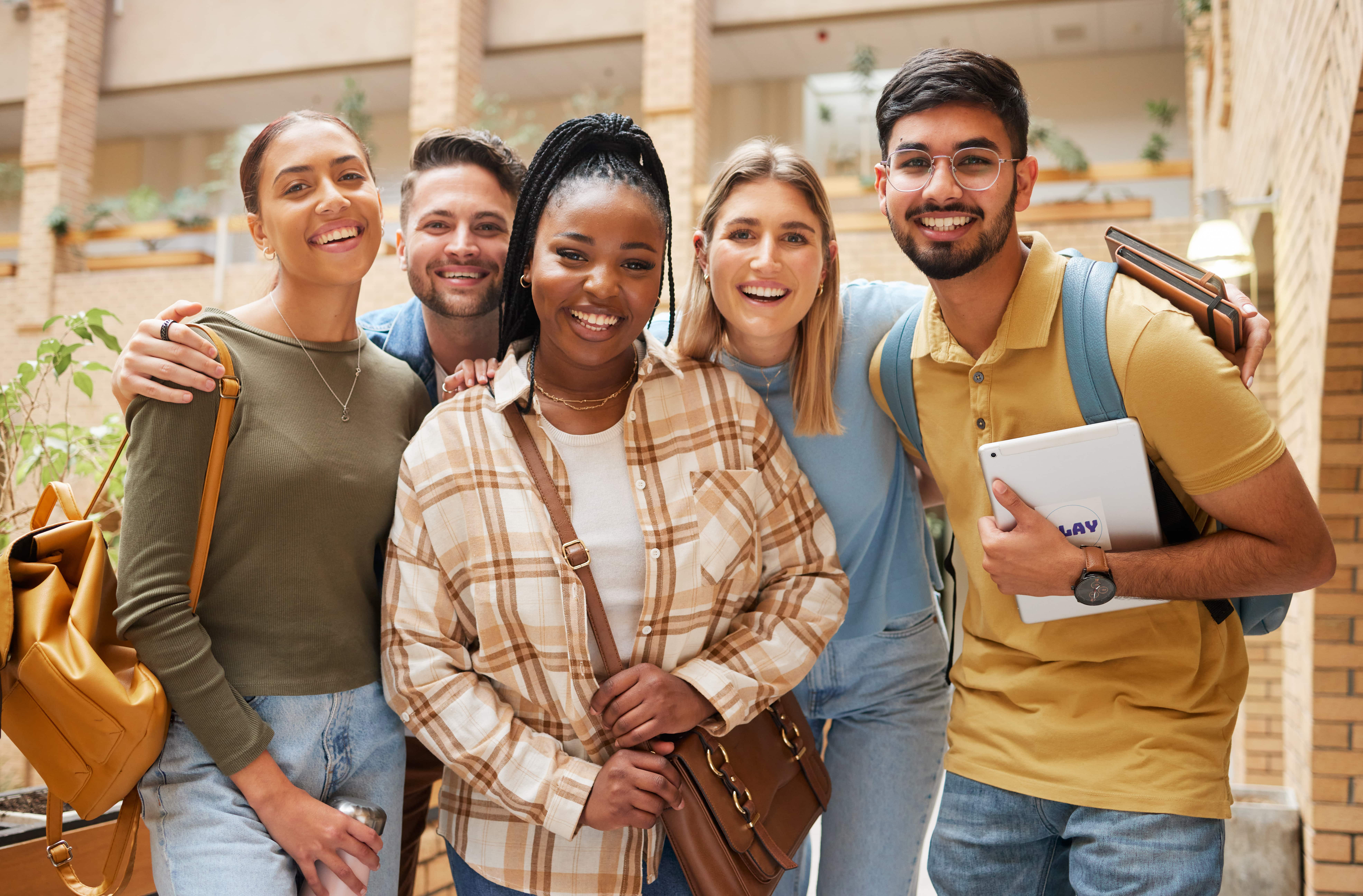 Grupo de cinco jovens sorridentes, de diferentes etnias, posando juntos em um ambiente escolar ou universitário. Eles usam roupas casuais e carregam mochilas e cadernos, transmitindo uma atmosfera de amizade, diversidade e ambiente acadêmico