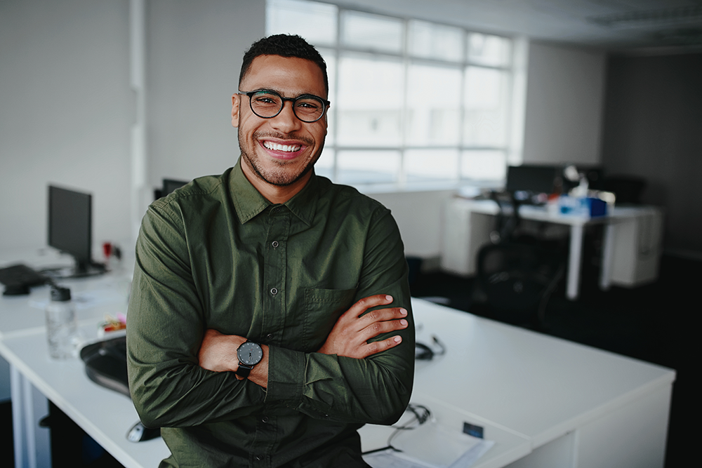 Jovem profissional sorridente, de braços cruzados, veste camisa verde escura e óculos, posando com confiança em um escritório moderno. A imagem transmite sucesso e preparo para atuar em diversas áreas de atuação da Administração, como gestão de pessoas, p
