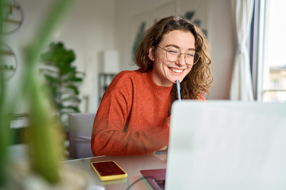 A imagem mostra uma jovem sorrindo enquanto trabalha no computador, possivelmente refletindo sobre como identificar seu perfil profissional em um ambiente claro e acolhedor. 