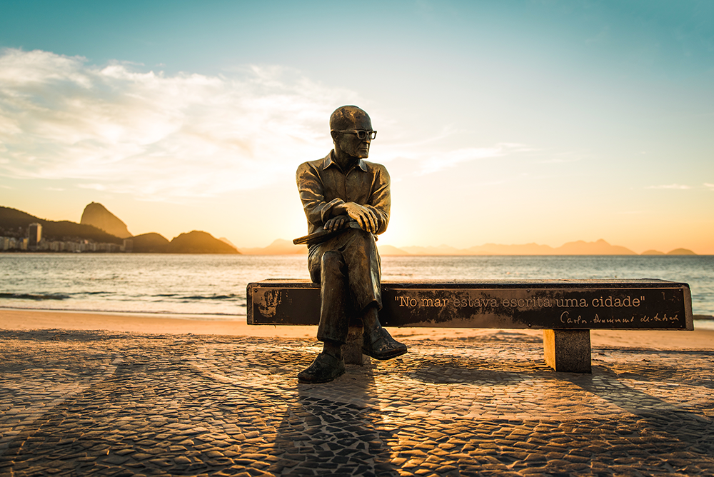 Uma foto da estatueta de Carlos Drummond de Andrade que fica no Rio de Janeiro, a praia ao fundo.