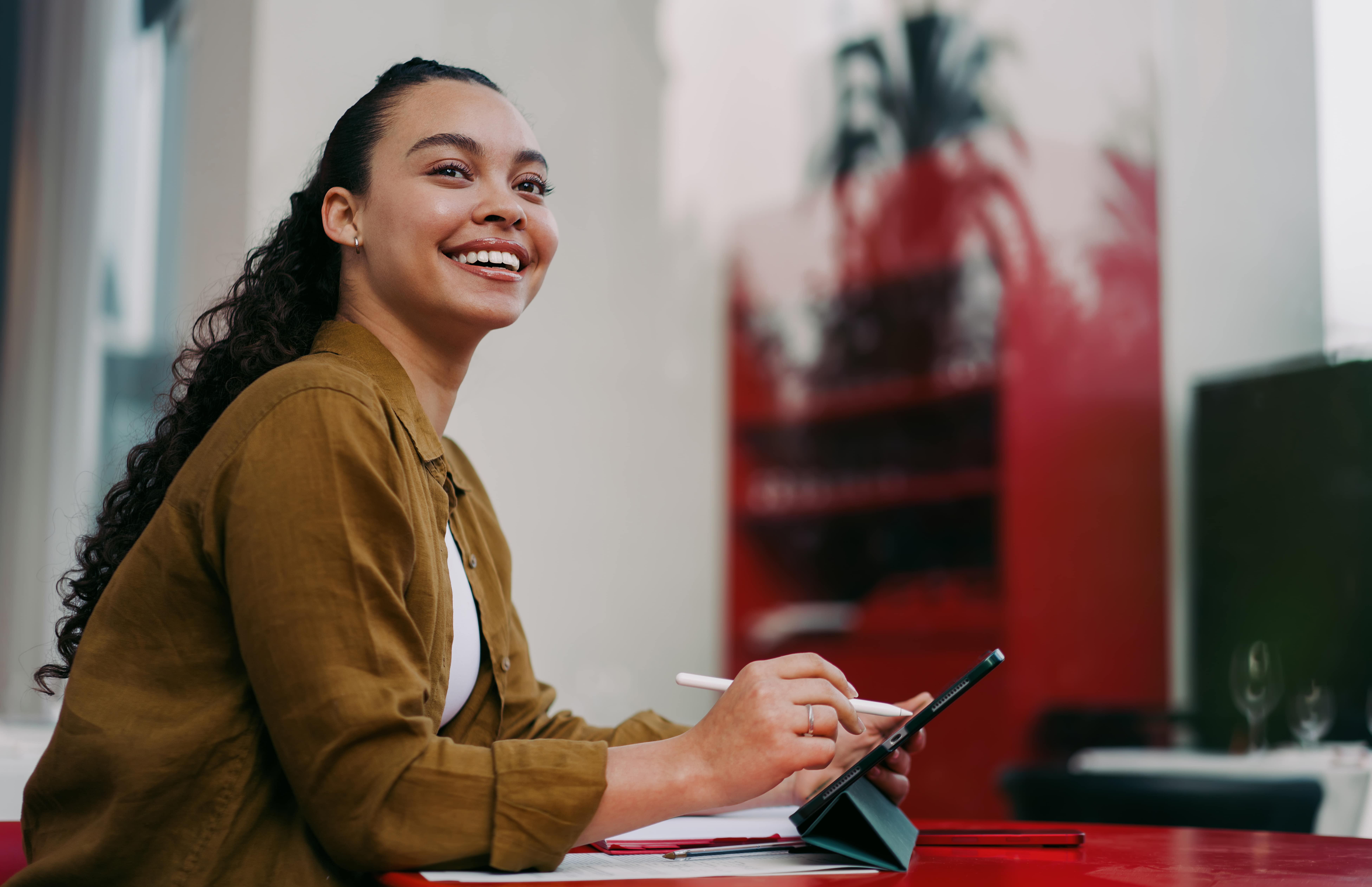jovem sorridente sentada em uma mesa vermelha, ao ar livre, usando um tablet com caneta digital. Representando a produtividade com IA em estudos e no trabalho.