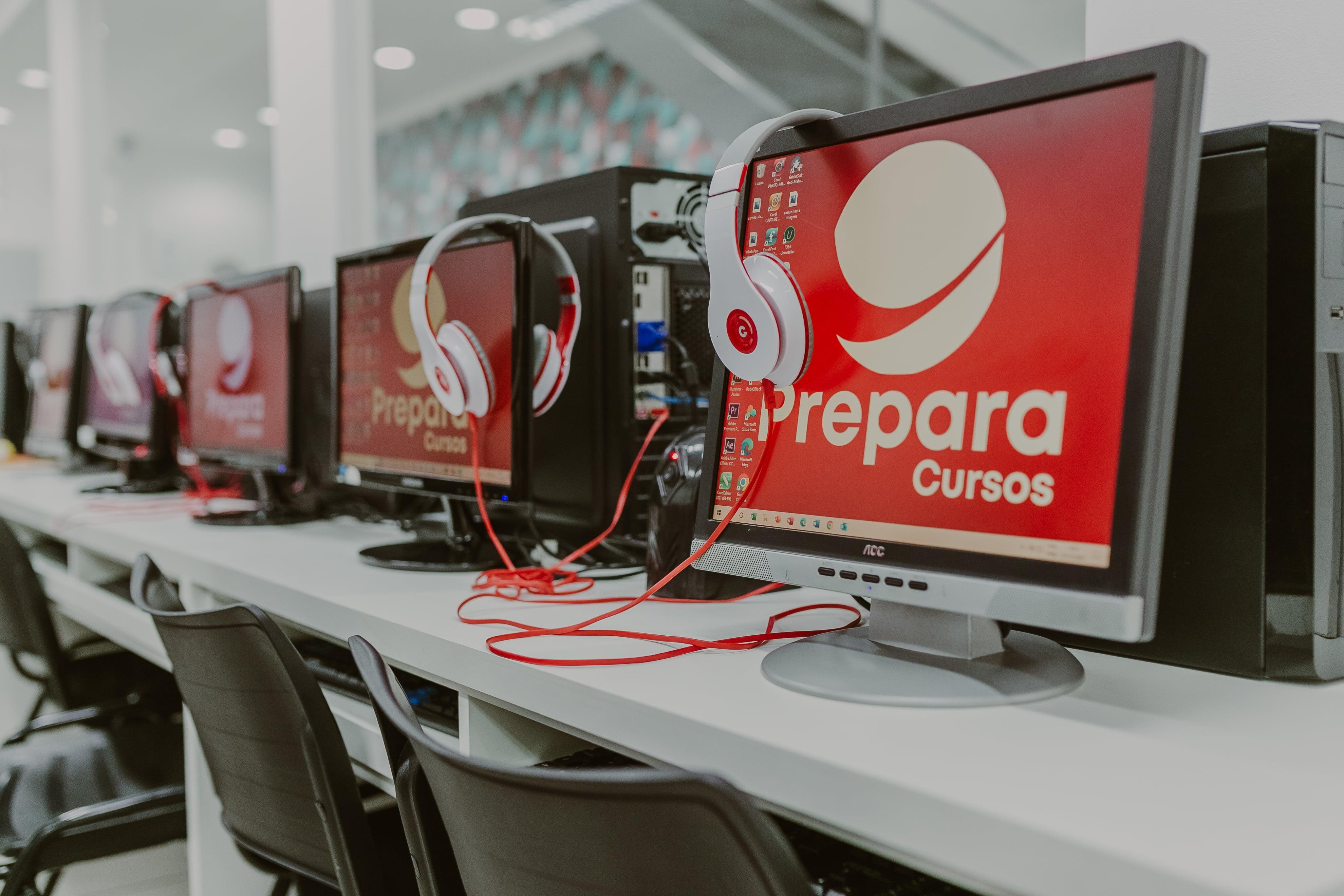 Fileira de computadores com fones de ouvido em um laboratório da Prepara Cursos, todos com a logo da instituição na tela, representando ambiente de aprendizado tecnológico e estruturado